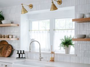 View of a kitchen sink under a bright window with handmade white tile on the walls and gold fixtures.