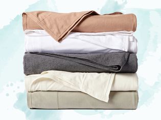 Five sets of the best jersey sheets in different colors stacked on a light blue watercolor background