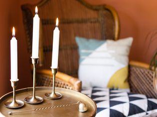 White candle sticks on brass candle holders in front of wicker chair with patterned pillows