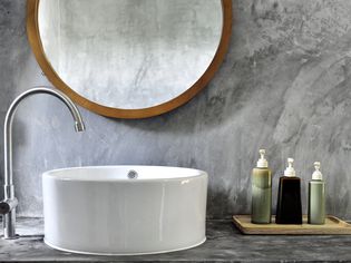 Concrete vanity with a white porcelain sink, a wood circular mirror, and soaps.