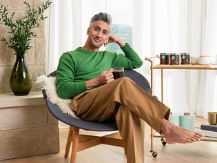Tan France poses in al iving room next to a bar cart with Starbucks coffee. He's holding a clear mug of coffee and wearing a green sweater and camel pants