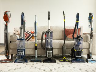 A side-by-side selection of seven stick Shark vacuum cleaners on rug in front of couch.