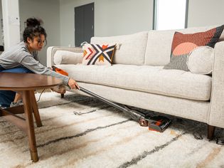 Woman using Shark HV301 Rocket Ultra-Light Corded Stick Vacuum to vacuum underneath a white couch sitting on a tan and white carpet